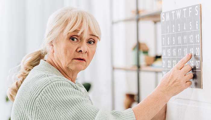 An older woman pointing at a wall calendar, with a concerned expression, in a softly lit home interior.